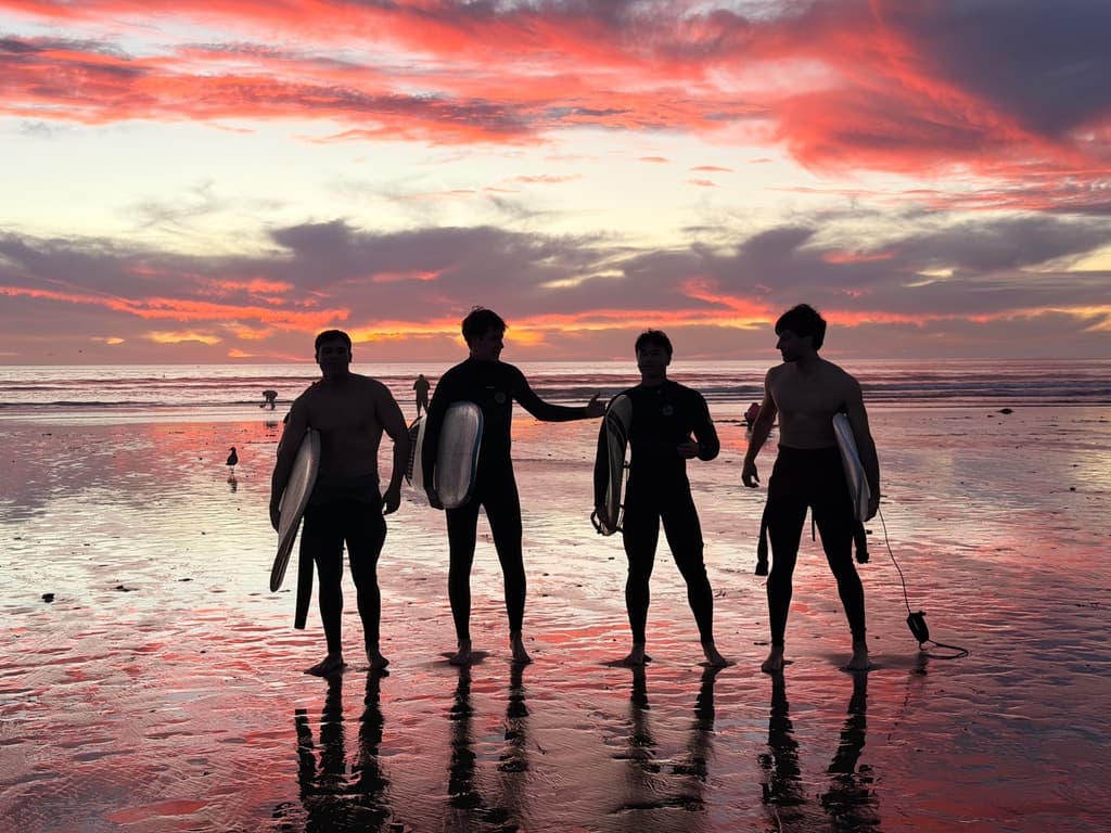 Four surfers silhouetted with their boards on wet sand at a red sunset
