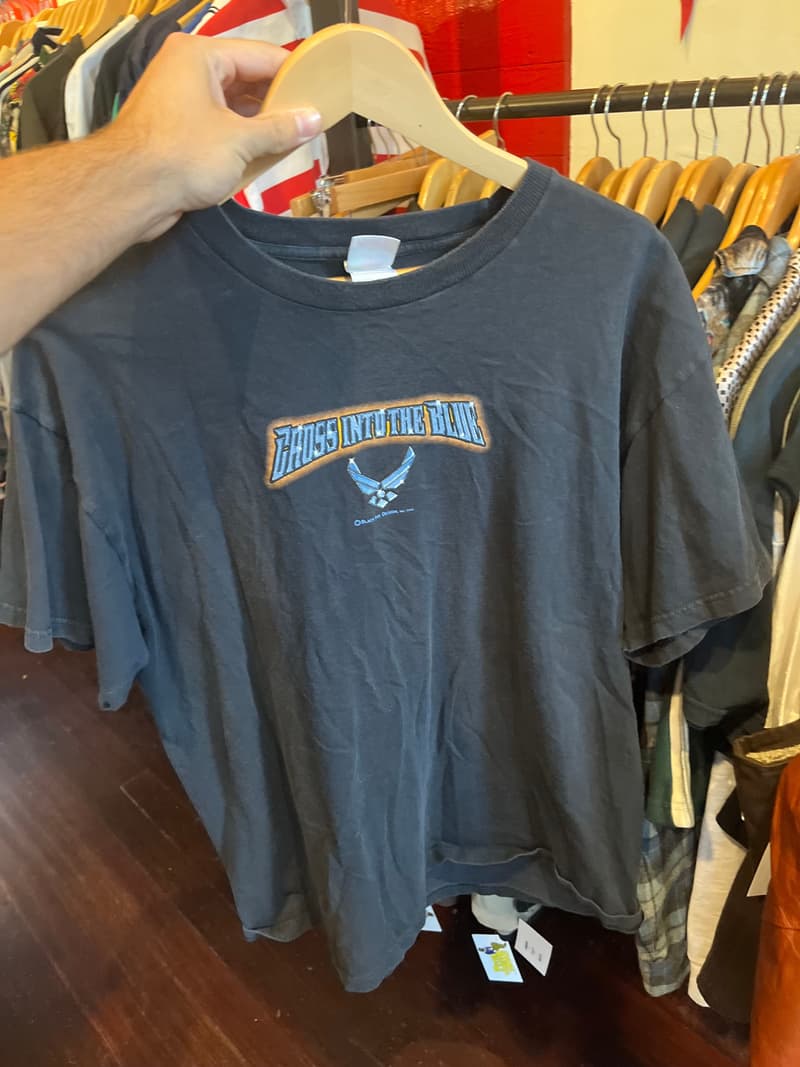 A faded gray Air Force graphic tee held up on a hanger in front of a thrift store rack